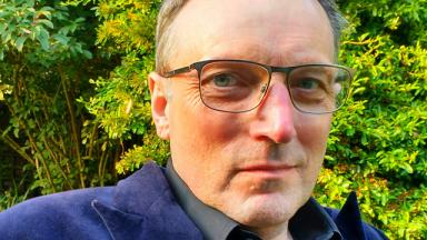 man with short hair and glasses cropped from shoulders up, green bushes in the background